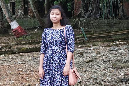 A young woman Ankita Bardhan standing in a purple and white printed dress in an open setting. The bottom of trees can be seen behind her