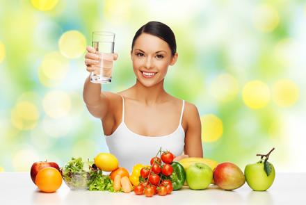 Stock pic of a young woman in a white tanktop behind a table of fruits and vegetables and holding up a glass of water