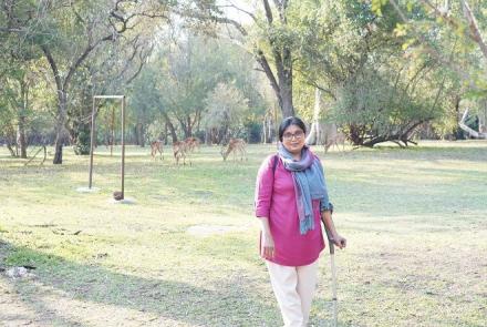 Neema in a pink shirt, white pants and a scarf in an open area walking with support of a cane 