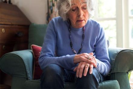Image: Stock pic of an older silver haired woman in a blue sweater sitting on a sofa and holding hands for an article on living well with Parkinsons Disease