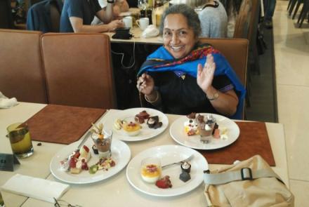 Image; Shaila Bhagwat, who has Parkinson's Disease in a blue dress at a restaurant with plates of desserts in front of her