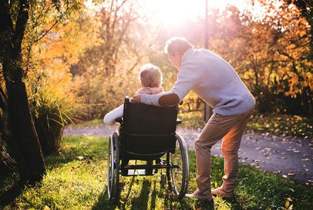 A couple, a woman on a wheel chair with a man standing attentively next to her, both looking into the woods with the sun streaming through