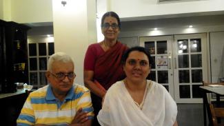Family pic inside a room of a young woman with a white dupatta seated with her silver haired father in a striped green and yellow t-shirt on the left and her mother standing behind them in a red sari and blouse