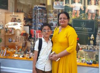 A woman in a yellow kurta standing with her young son in a white shirt in front of a shop window. Both wear spectacles.