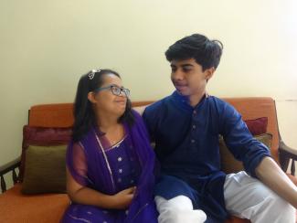 Aarshia on the left a young Indian black haired spectacled girl with Down Syndrome in a purple dress and on the right her brother Aaryamann a young Indian teen boy with black hair in a purple kurta and white pyjamas