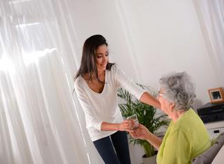 Stock pic of a younger person in a white top and black pant supporting and holding hands with a seated silver haired elderly lady in a green top