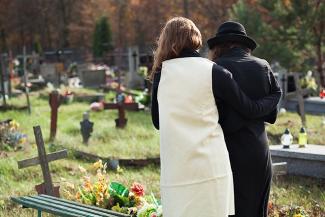 Image: Backs of woman in a white dress with a man in a black suit and black hat in a cemetery mourning passing of a loved one