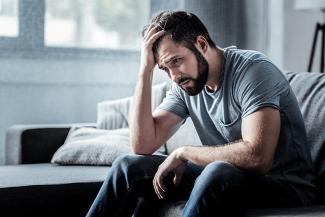 A man in grey shirt and jeans sitting on a couch and holding his head in despair