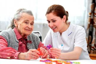 A caregiver in a white dress does an activity with an elderly person with dementia in red and grey 