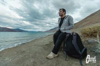 TPic of the author in a grey jacket, black t-shirt and black track pants, resting on a rock in front of a water body