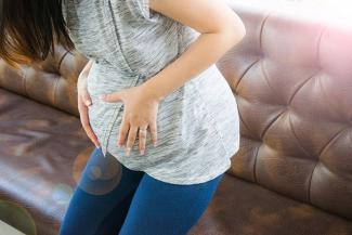 Image: A visibly pregnant woman in blue pants and a light top holding her baby bump 