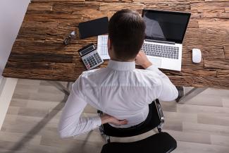 Stock pic of the back of a man feeling rheumatic pain while working at his desk on a laptop