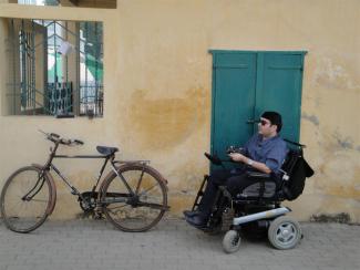 Rustom Irani on the right in a wheelchair in front of a blue door and on the left is a wheelchair against a wall 