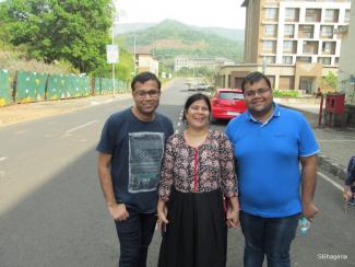 A mom in a printed top and black skirt flanked by her adult sons,  the one on the left with a dark t-shirt and the one on the right with a blue t-shirt. In the background is a red car parked in front of a building on the right and a fence and trees on the left