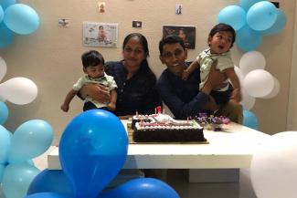 A couple with their kids on either side with a cake on the table in front and blue and white balloons around 