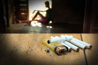 Image of a person sitting in a doorway and unused cigarettes on the table in the foreground depicting smoking or nicotine withdrawal