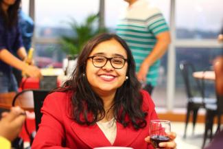 A young woman, spectacled, shoulder length hair in a red dress in the foreground and holding a glass of wine. In the background are a few tables and blurred images of 2 people in blue shirt and green tshirt pulling chairs 