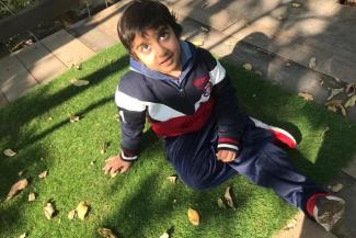 A young boy in navy blue pants, and blue, red and white jacket and red and white shoes sitting on the green grass and looking up at the camera