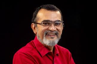 A profile pic of a smiling Ganesh N. Rajan with spectacles and a black and white peppered beard in a red shirt with black background