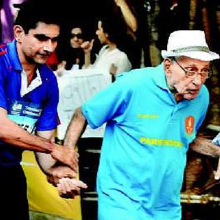Parkinsons patient Merwan Zend in front in a light blue shirt and hat walking in front supported by another young man in a darker blue shirt 