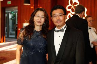 On the left is Yee Ling with her husband Yen-Lu Chow on the right. She is dressed in a black formal dress and he is wearing a black tuxedo with a white shirt. The background is red and shows some people blurred 