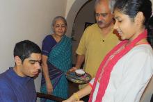 Image Description: Nivriti on the right in a white kurta and red dupatta tying a rakhi on her autistic brother Nishant's wrist. Nishant is sitting and wearing a blue kurta. Looking over both of them is their father in a mustard kurta and grandmother in a blue sari