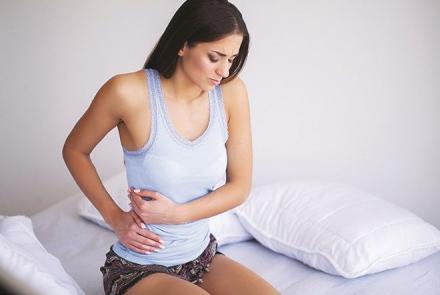 A woman wearing a tank top and sitting on the bed clutching her side