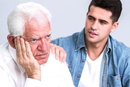 Image of an older silver haired person thinking and a younger dark haired person in a blue shirt and white t-shirt sitting next to him and debating whether to share or not about Parkinson's Disease diagnosis
