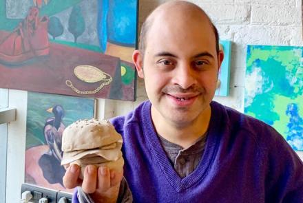 Shaurya a young man with down syndrome in a blue shirt in front of his paintings holding a burger he sculpted