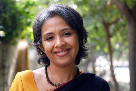 Head shot of the author Gayathri Prabhu wearing a yellow blouse, brown sari and a necklace around her neck. In the background are trees 