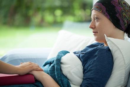 A stock pic of a woman as a patient resting wearing a bandana on her head and her hand is being held by another person who is not visible in the pic