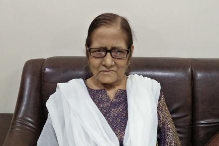 A bespectacled woman in a brown dress and white dupatta sitting on a sofa 