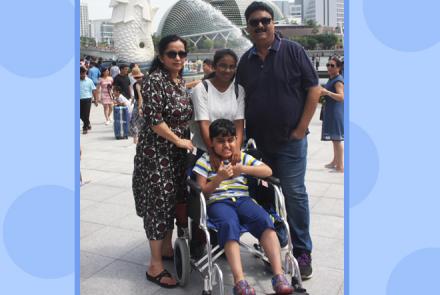 Rishi Chowhan a young child with Duchenne Muscular Dystrophy and Autism on a wheelchair with his family standing behind him from Left to Right- his mother, his sister and his father. Picture shows Singapore's Merlion in the background 