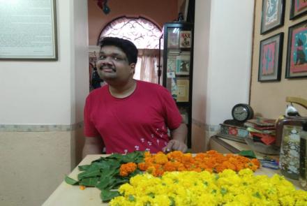 Kabir Kumtha, a young man with autism wearing a red tshirt standing in front of a table and working with yellow and orange marigold or jhendu, his favourite flower to make torans or garlands