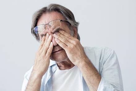 Stock pic of a grey haired man in a light shirt over a white undershirt pressing down his eyes in pain with his specs pushed onto his forehead