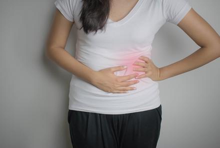 Stock pic of a woman in a white t-shirt and black pants holding a flare up in her abdomen area demonstrating PCOS