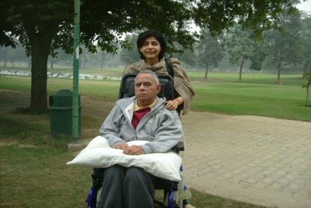 Rima standing behind her father who is in a wheelchair and has a pillow on his lap