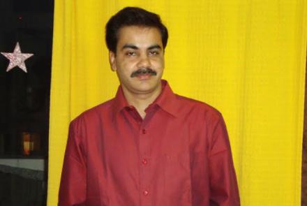 Image: Sanjay Jaiswal, with black hair and a moustache is an Oral Cancer survivor and stands in a red shirt against a yellow background