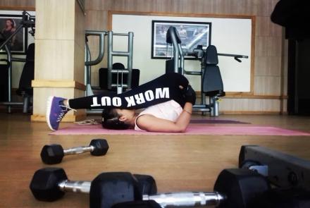 A woman in a yogasana at the gym. In the foreground are dumbbells, in the background gym equipment