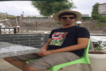Tanmay is a young adult on the autism spectrum and is wearing a black tshirt and a hat  and sunglasses and sitting outdoors on a green chair