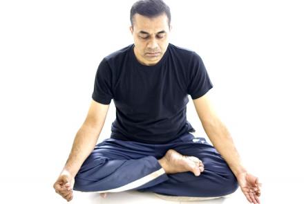 A man in a black t-shirt and blue yoga pants sitting in padmasana or lotus pose