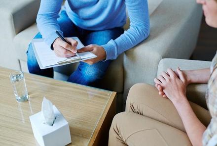 A stock pic of partially visible person sitting in a consult session with a partially visible counsellor with a note book in her hand. In front of them is a table with a glass of water and a box of tissues