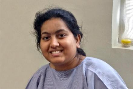 A smiling black haired young woman with Lupus in a mauve coloured hospital gown