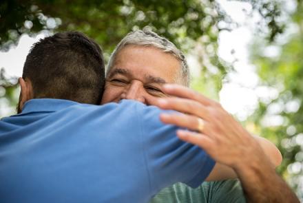 A view of two people hugging each other under a tree, the back of one in a blue shirt and the face of the other is visible