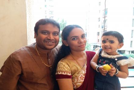 Person with haemophilia, Vinay Nair in a brown kurta with his wife in a red and gold sari holding baby girl in green and white dress in arms